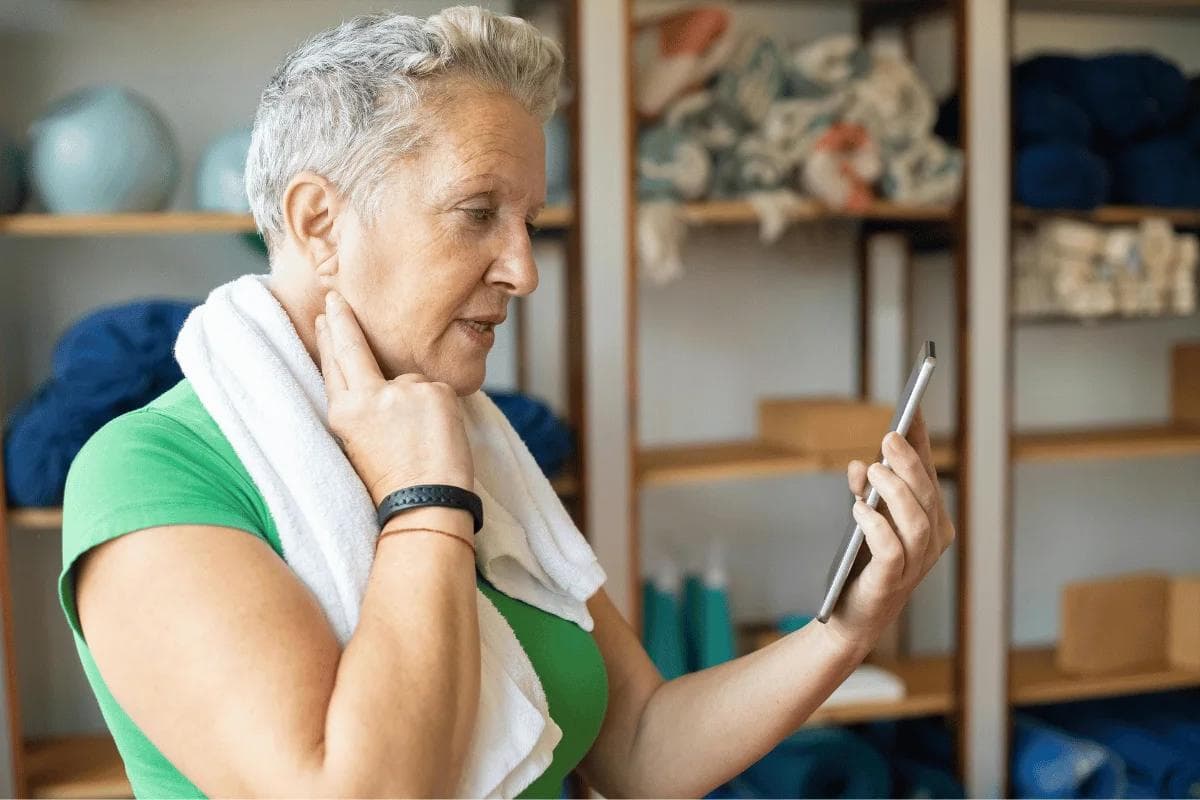 Older woman in gym attire using smartphone while holding her neck after workout.
