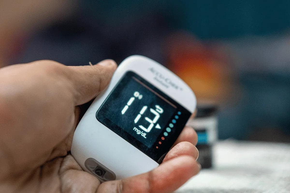 A hand holds a digital glucometer displaying a blood sugar level of 113 mg/dL, set against a blurred background, indicating health monitoring.