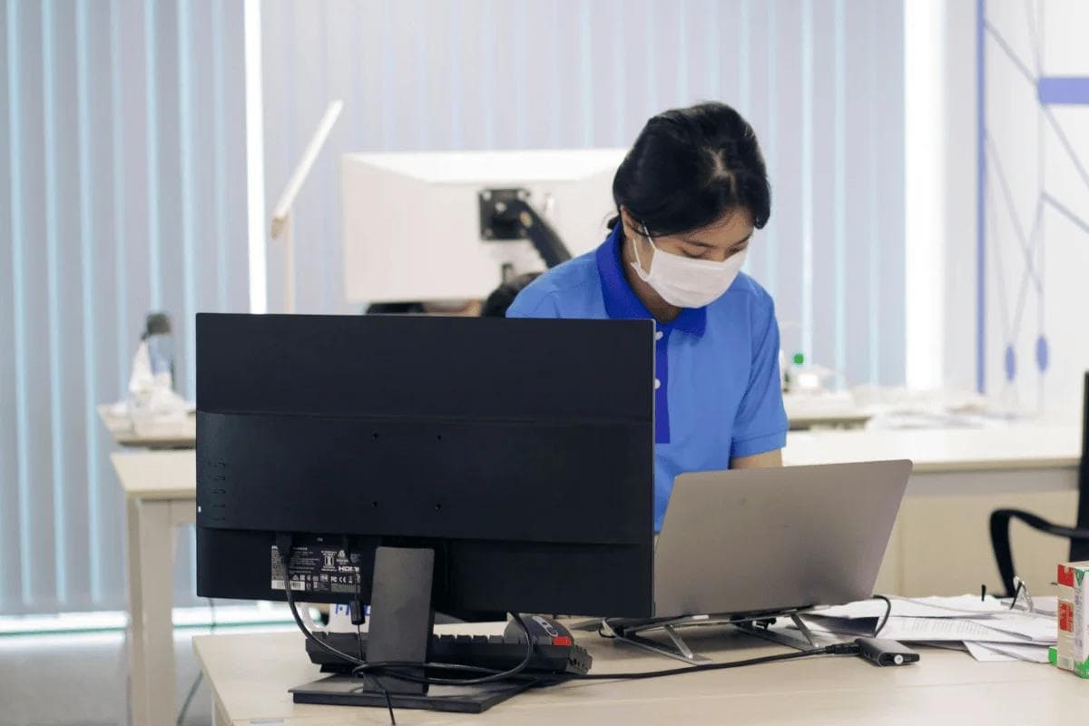 Office worker wearing a mask while working on laptop and monitor in modern office.