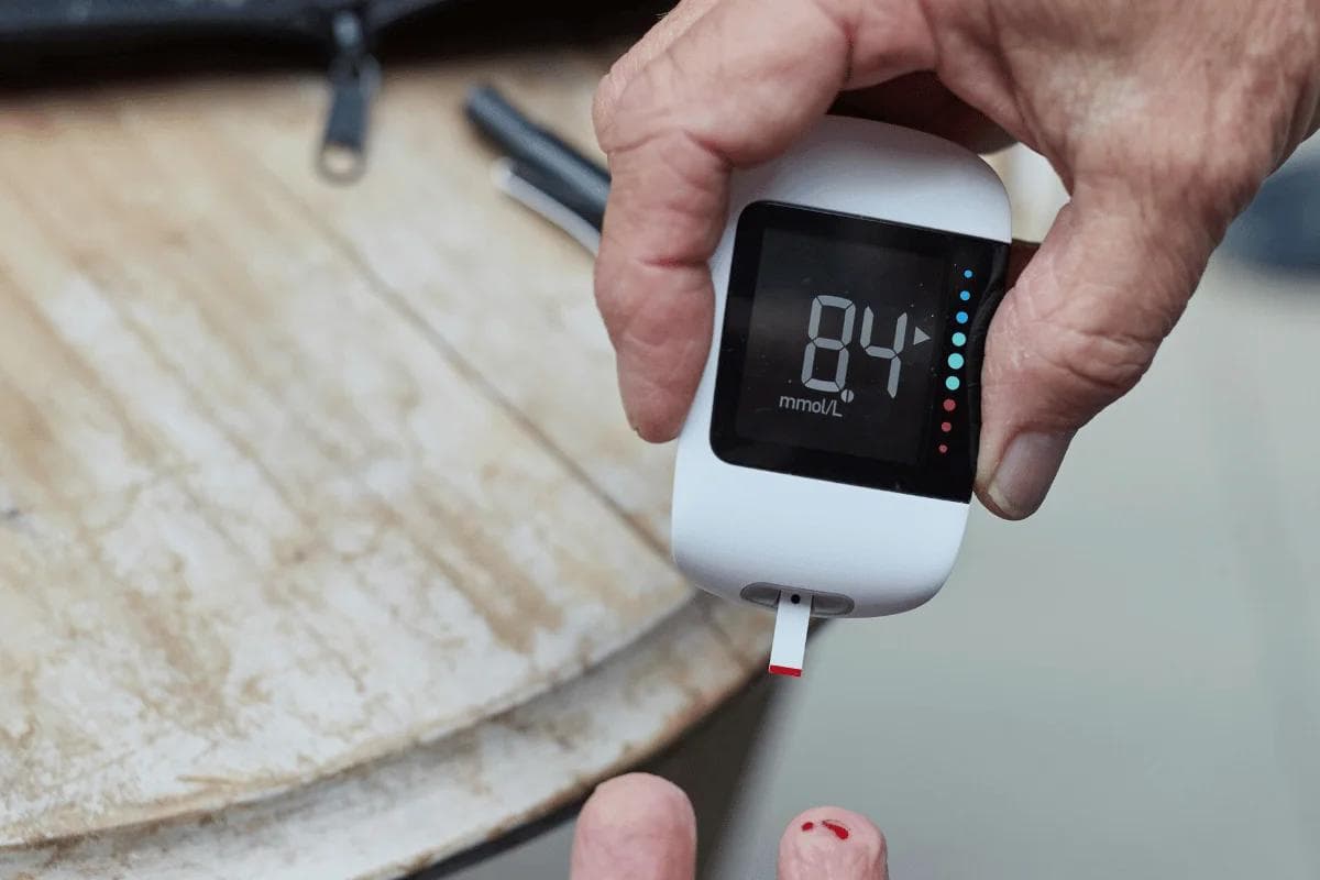 A hand holds a glucose meter displaying a reading of 84 mmol/L, with a blood sample on the fingertip and a blurred background.